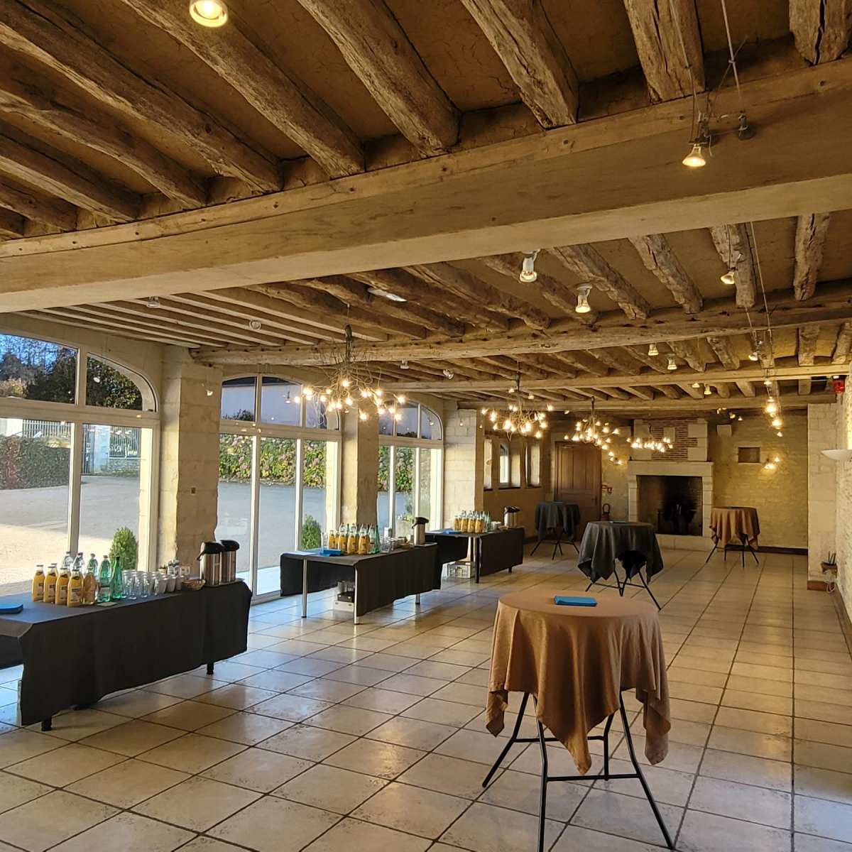 A spacious event room with wooden beams, featuring several tables with beverages and light fixtures, and large glass doors leading to an outdoor area.