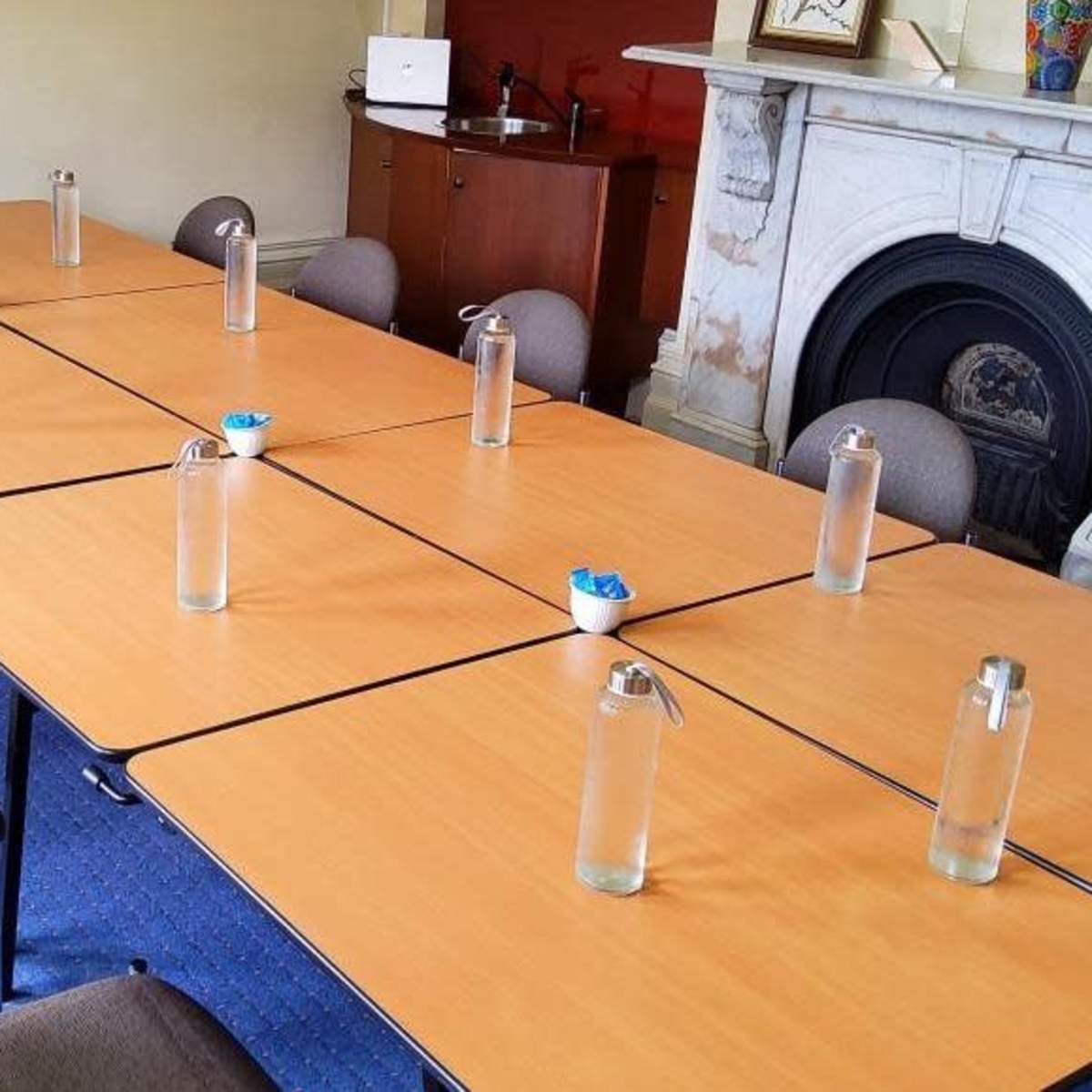A meeting room is set up with several tables arranged in a U-shape, each containing a glass water bottle and chairs positioned around them.