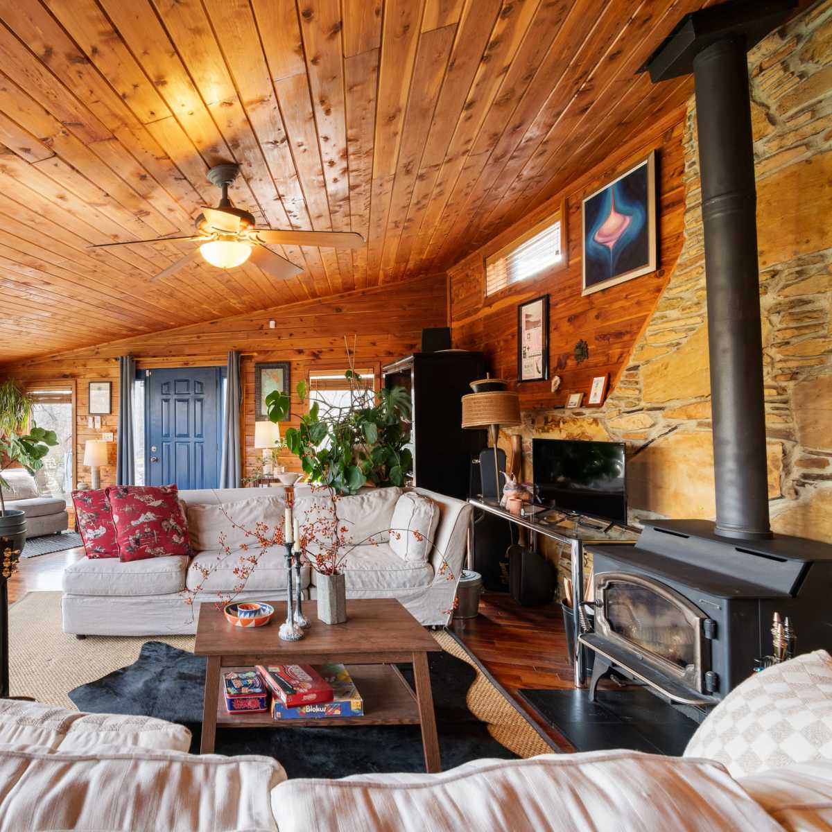 The living room features a wooden ceiling, two large couches, a coffee table with decor, and a stone wall with a wood stove, all surrounded by windows that offer views of the outdoors.