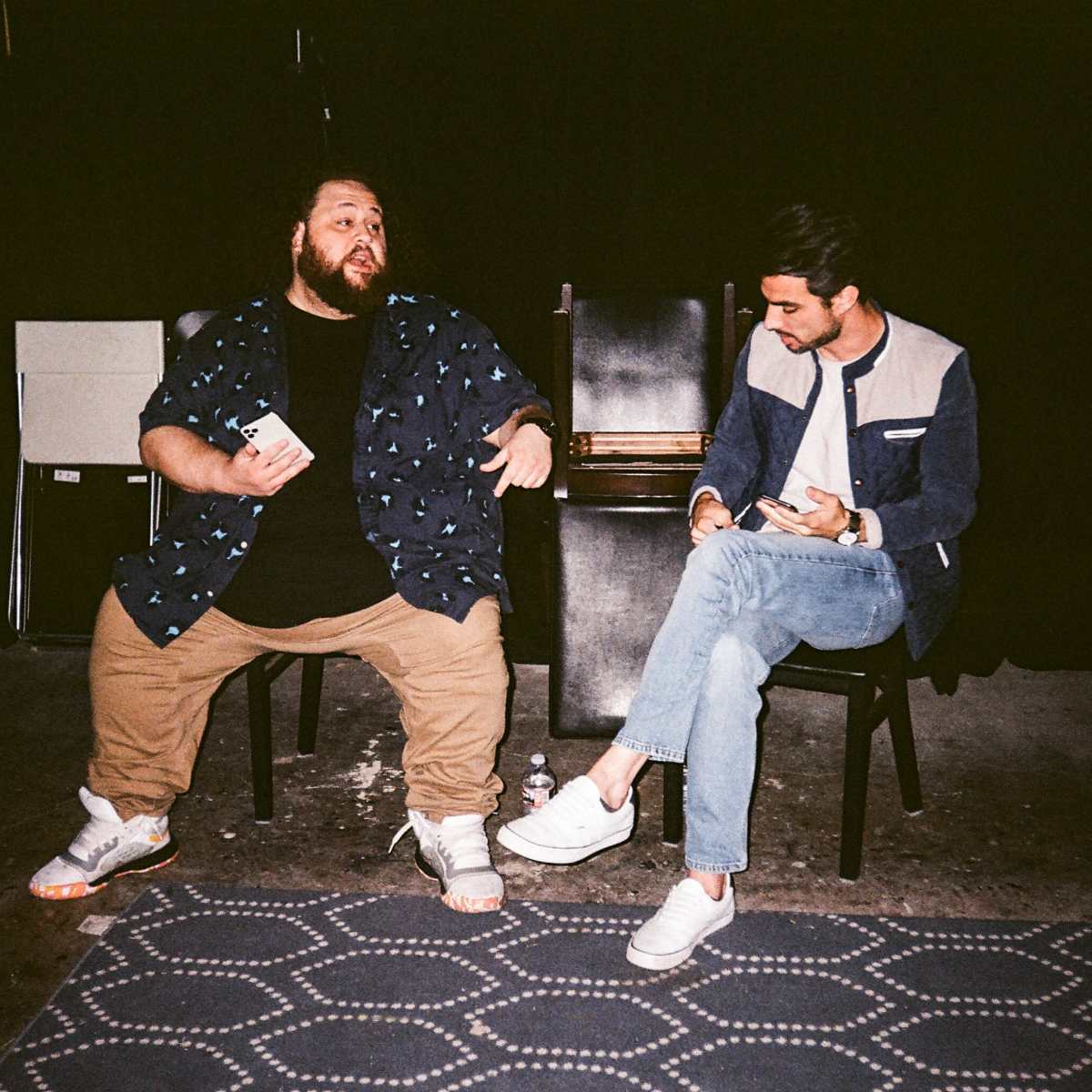 Two men sit on chairs in a dimly lit space, engaged in conversation, with one holding a small item and the other looking at his phone.