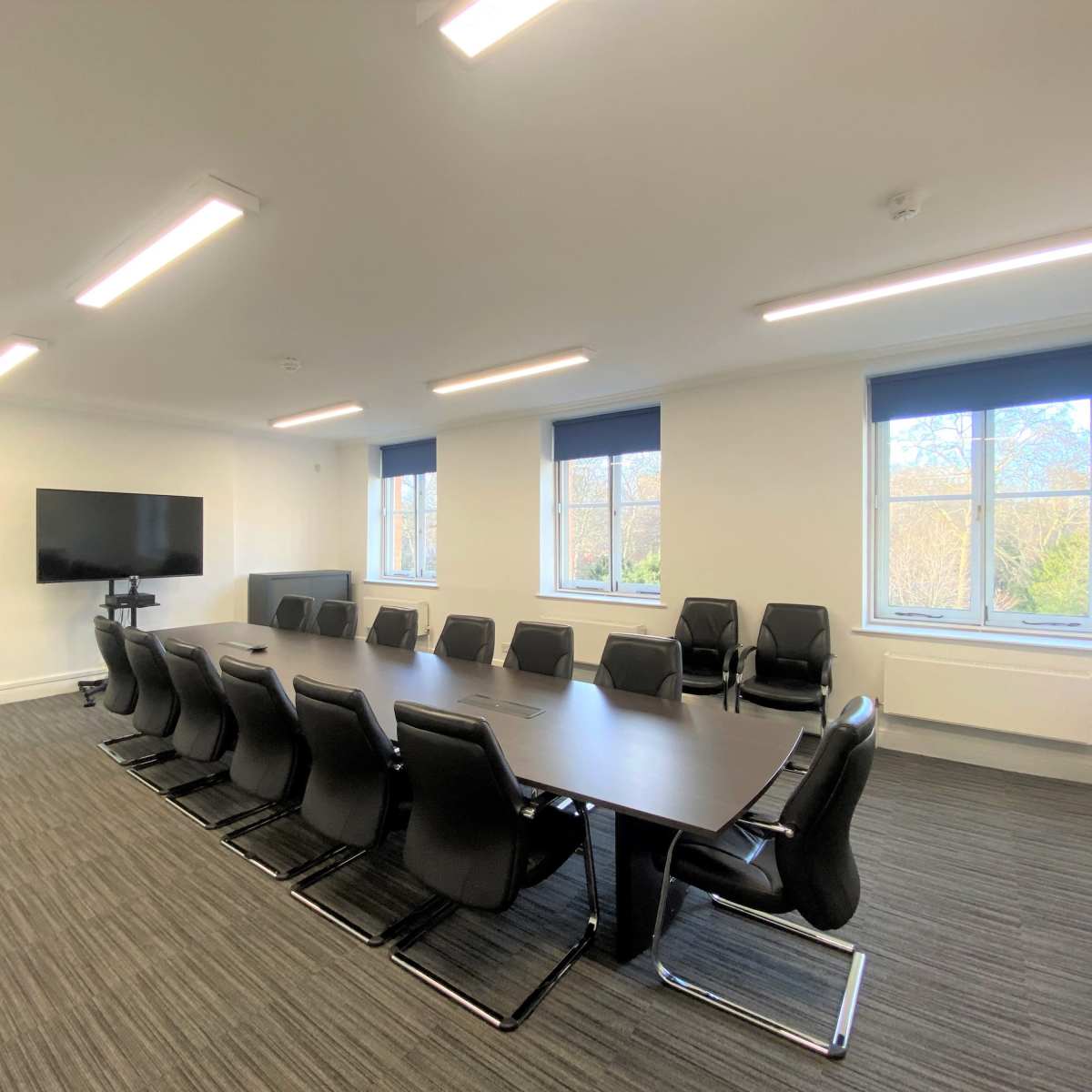 A modern conference room with a long table, black chairs, a large screen on the wall, and windows providing natural light.