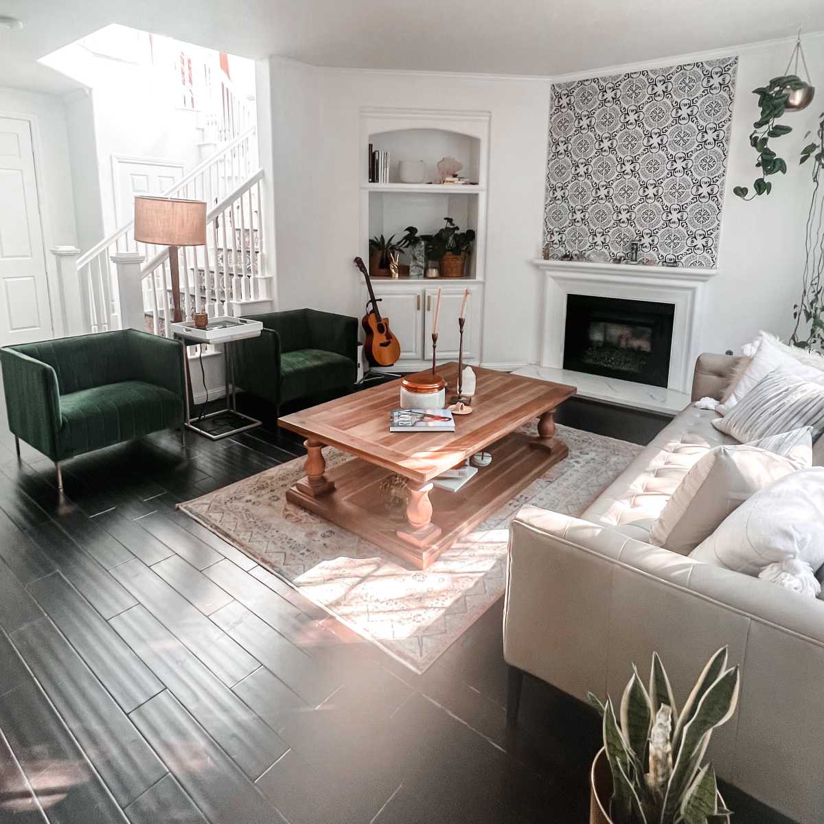A bright living room with a cream sofa, green armchairs, a wooden coffee table, and a guitar placed near a white fireplace.