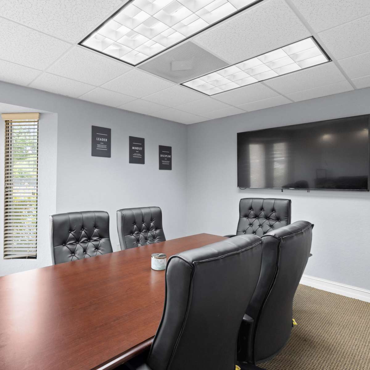 A conference room features a wooden table surrounded by black leather chairs, with blinds covering the windows and a wall-mounted television.