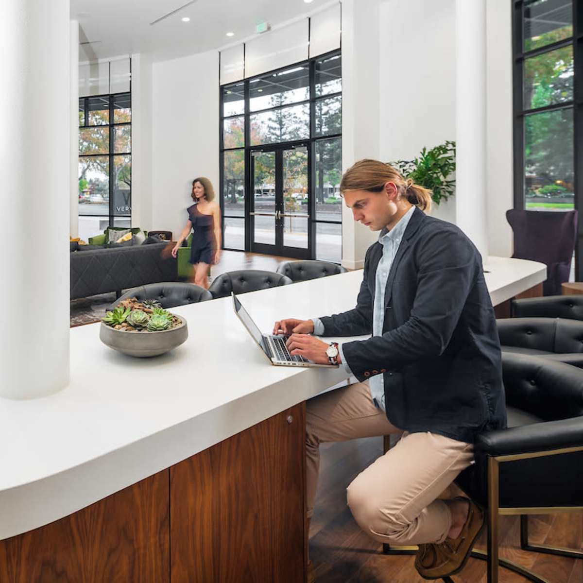 A man sits at a curved white countertop using a laptop while a woman walks through a modern lobby with large windows and contemporary furniture.