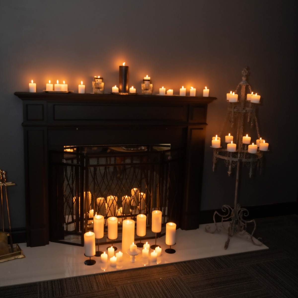 A dark room features a fireplace adorned with numerous glowing candles of varying heights, creating a warm illumination.