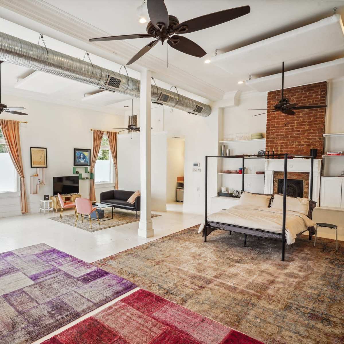 A spacious, modern room with exposed brick, large windows, two ceiling fans, and a mix of seating areas and a bed.