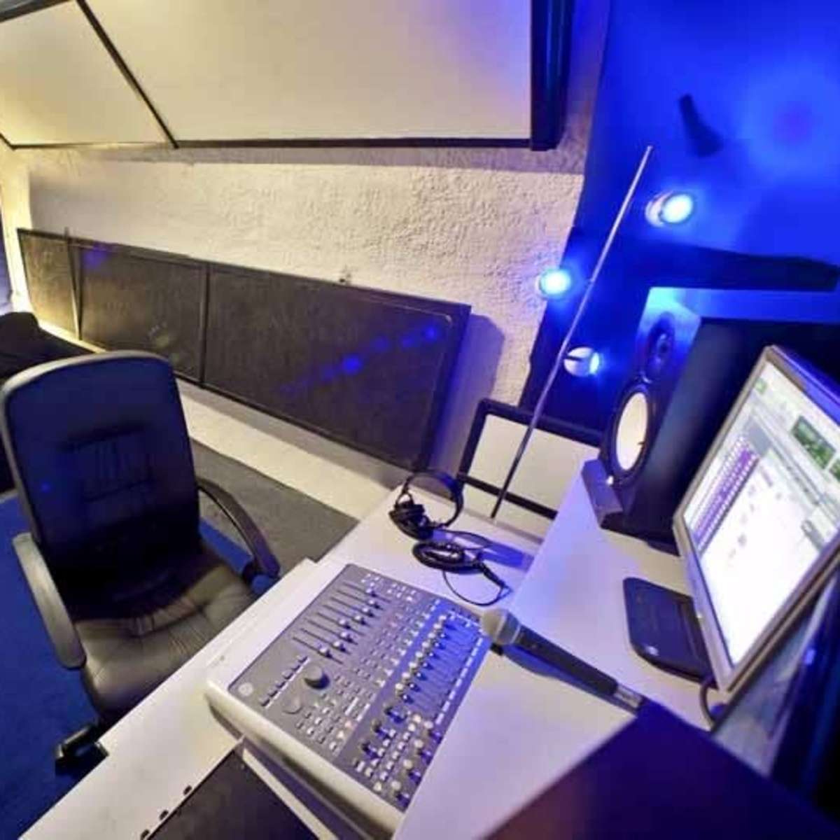 A compact studio space with a desk and computer setup, a black bed, and a chair positioned on a blue rug.