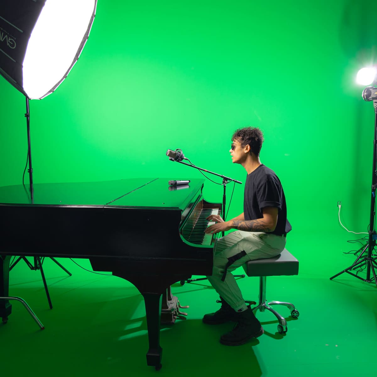 A person sits at a grand piano in front of a green screen, with studio lights positioned nearby.