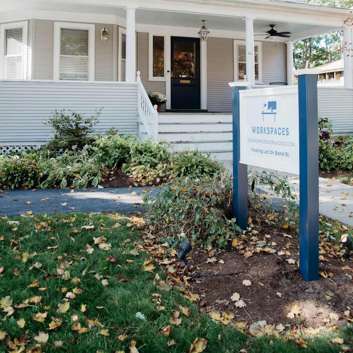 A light-colored, two-story house with a front porch and a sign advertising workspaces stands in a landscaped area with autumn leaves on the ground.