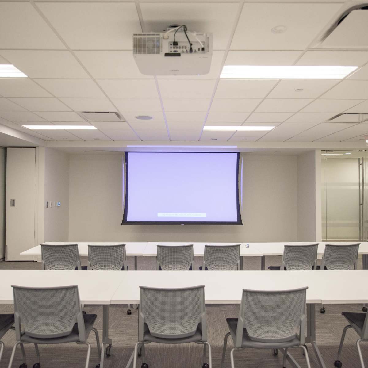 A modern conference room features rows of chairs arranged in front of a large screen and white tables.