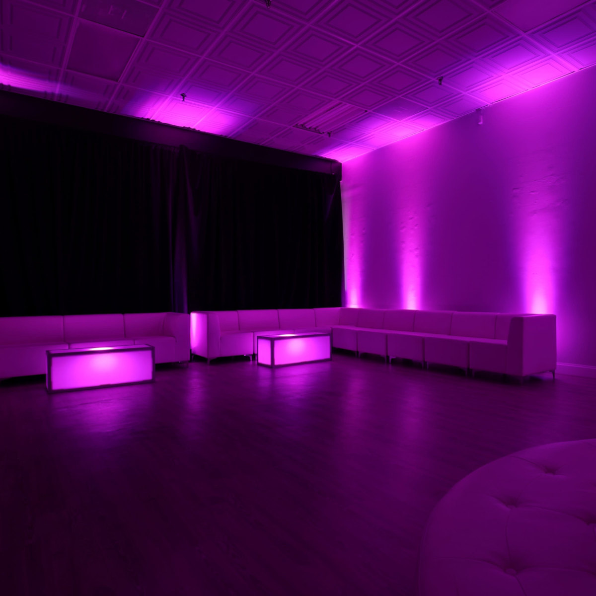 A modern lounge space with purple lighting, featuring white seating and illuminated coffee tables.