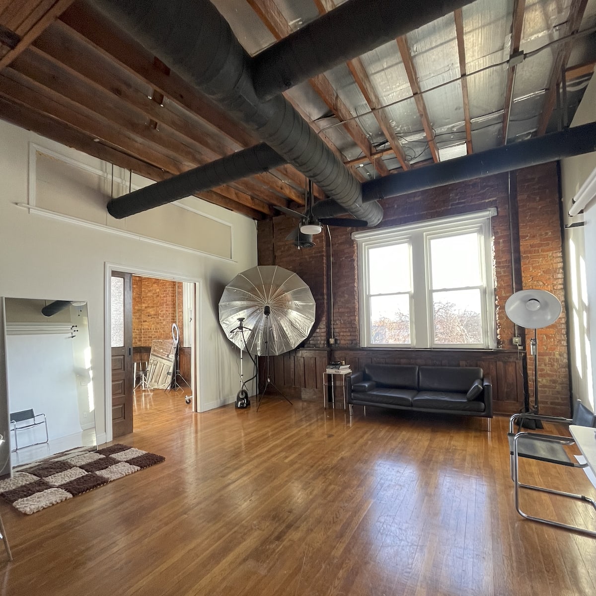 A spacious room with wooden floors, exposed brick walls, and large windows, featuring a black sofa, a large silver reflector, and a few pieces of minimalist furniture.