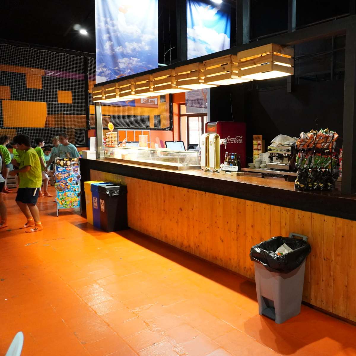 A brightly lit concession area with a wooden counter and various snack and drink options displayed, along with two trash bins nearby.