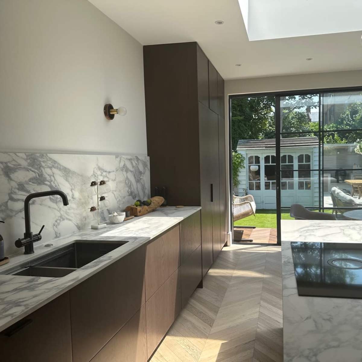 A modern kitchen features marble countertops, dark cabinetry, and a view of a garden through large glass doors.