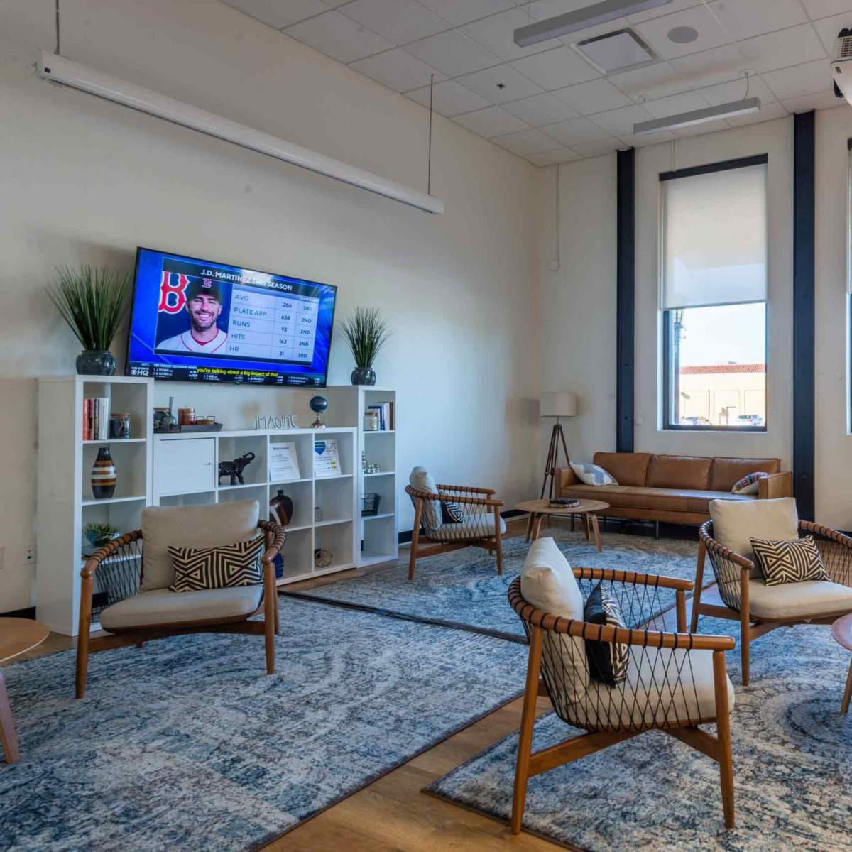 A modern lounge area features comfortable seating, a television displaying sports content, and large windows letting in natural light.