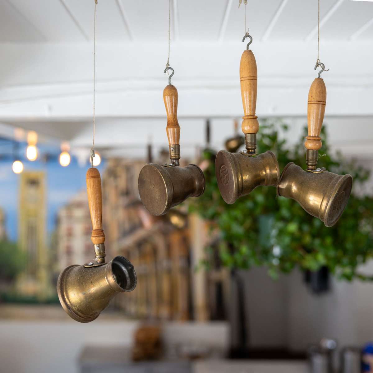 Brass coffee pots with wooden handles hang from a white ceiling in a café decorated with a city mural.