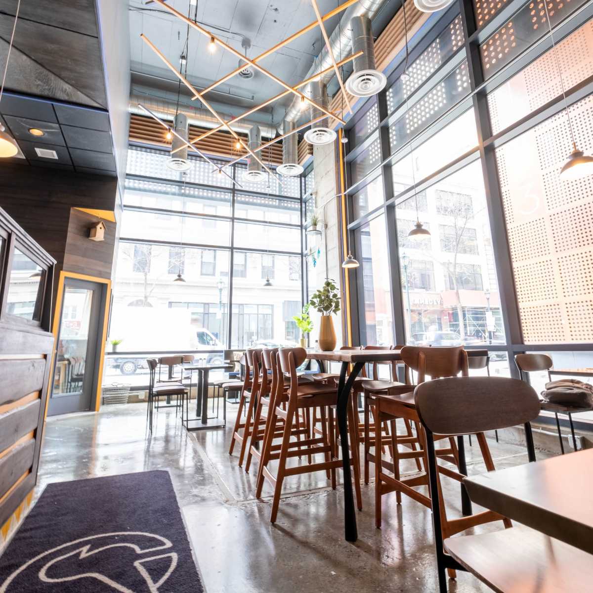 A modern café interior featuring wooden furniture, large windows, and decorative ceiling lights.