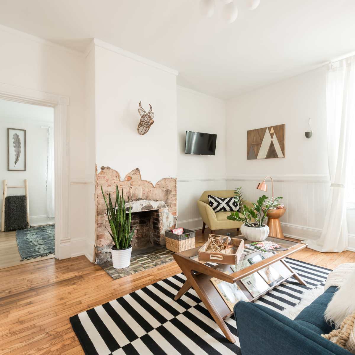 A living room features a mix of furniture with a coffee table, a television mounted on the wall, and large windows allowing natural light to illuminate the space.