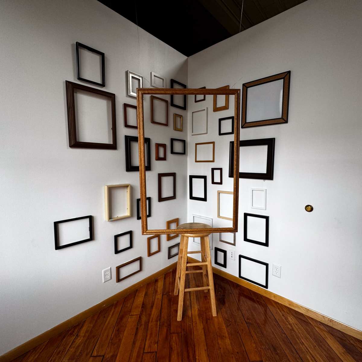 A large wooden frame stands on a stool against a wall adorned with various sizes and styles of empty picture frames.