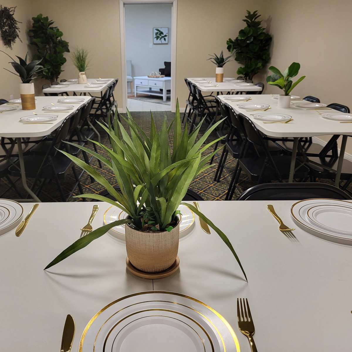 A neatly arranged dining area features several tables set with plates, utensils, and potted plants at the center.