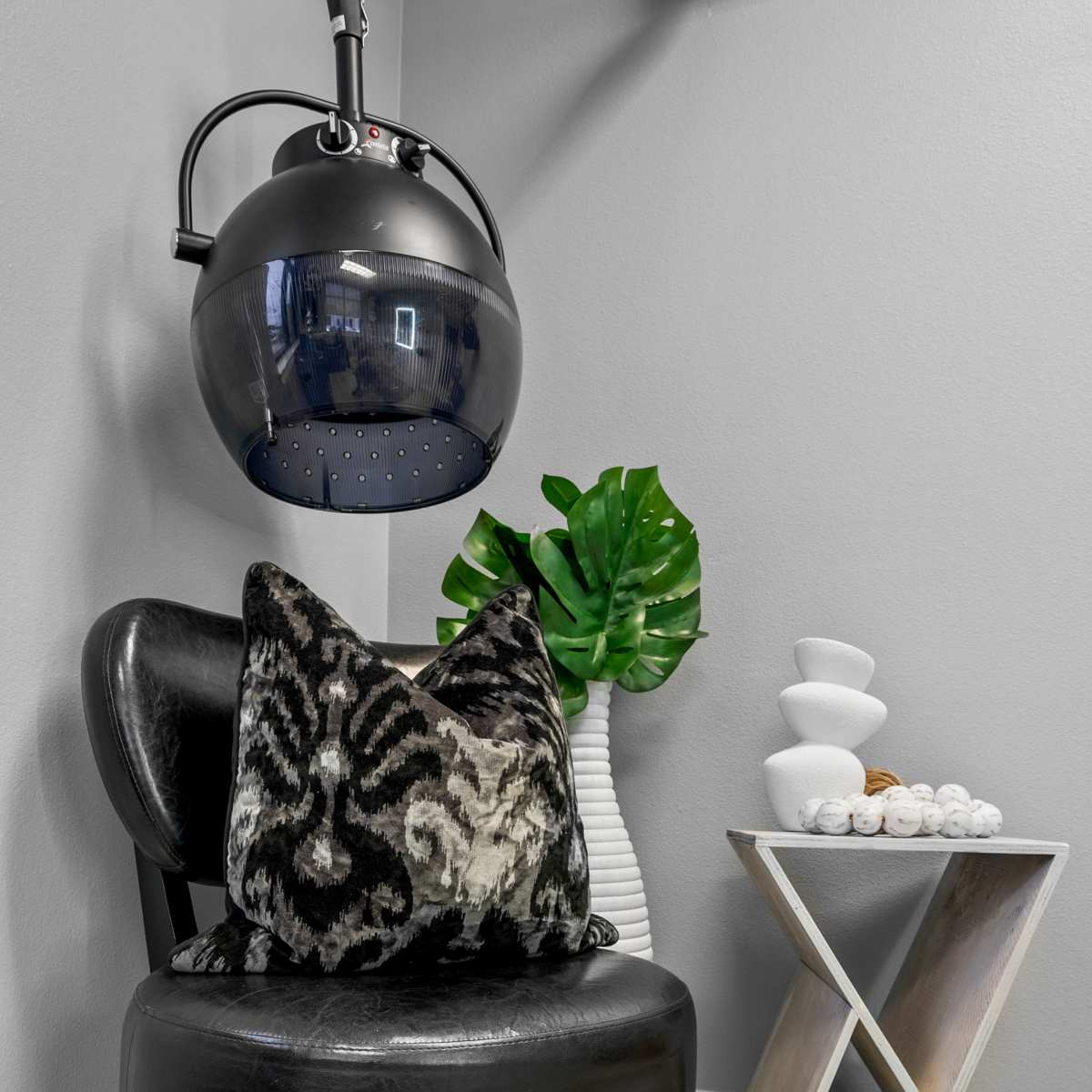 A black chair with a patterned pillow sits next to a decorative plant and a modern side table in a well-lit room.
