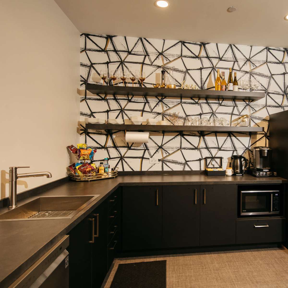 A modern kitchen with black cabinets, a stainless steel refrigerator, and open shelves displaying glassware and kitchen items against a patterned wall.