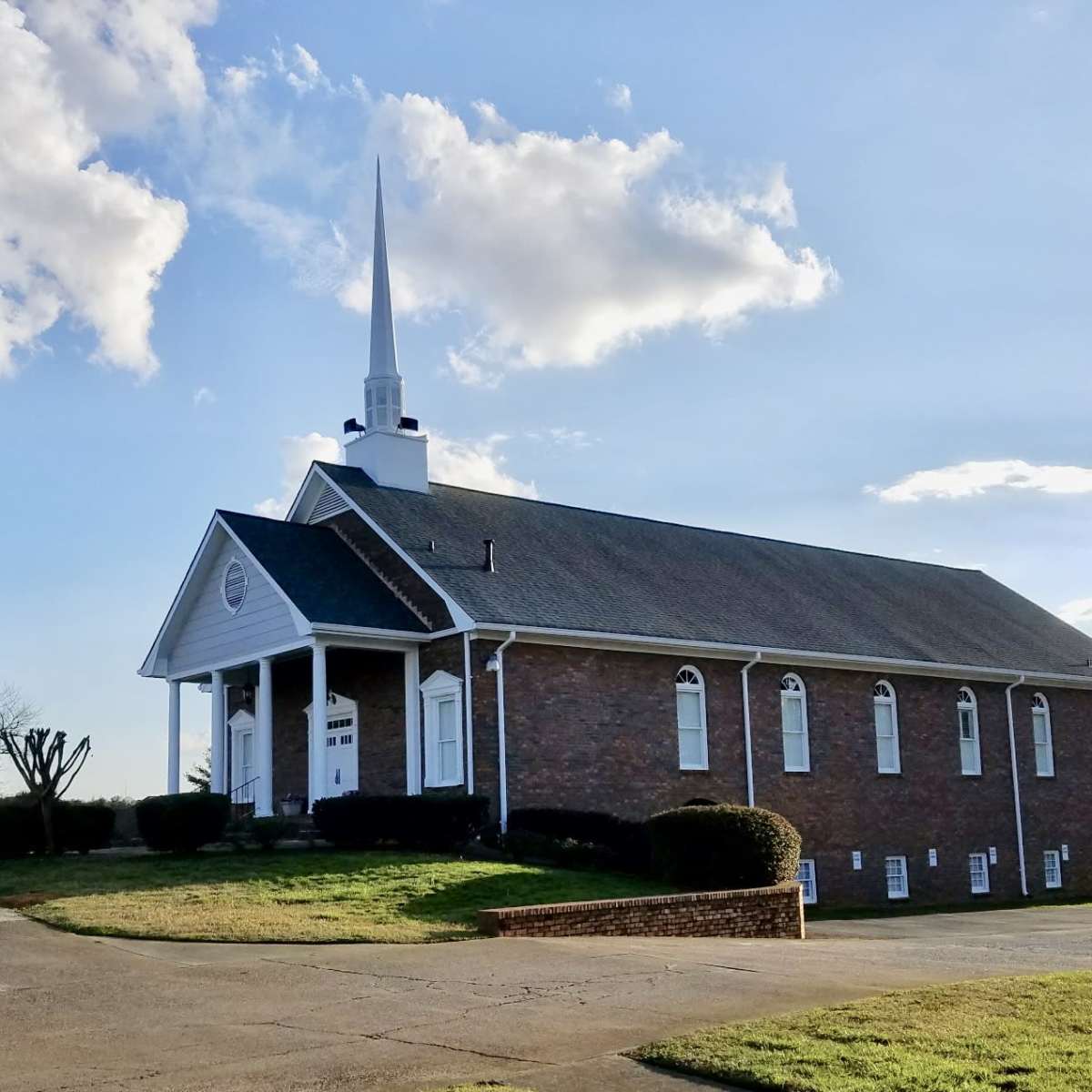 A brick church building with a steeple stands at the end of a paved driveway under a blue sky with clouds.