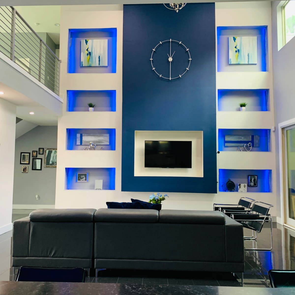 The interior of a modern living room features a dark gray sectional sofa facing a wall with blue backlighting, a clock centerpiece, and a mounted television.