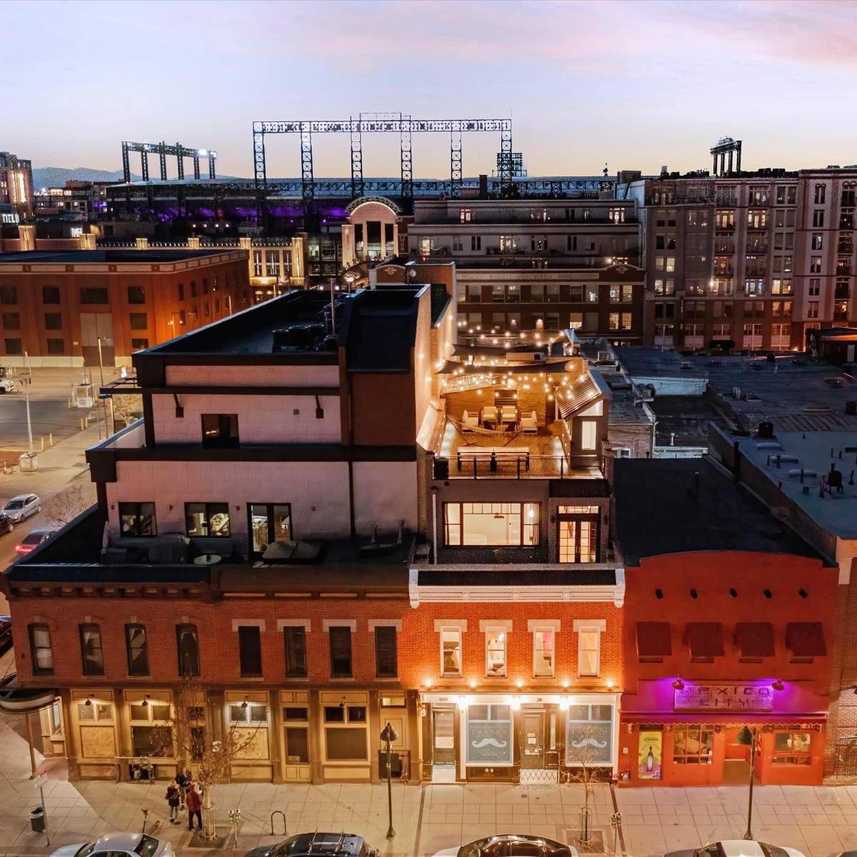 A rooftop restaurant atop a multi-story brick building in an urban area during twilight, with city lights visible in the background.