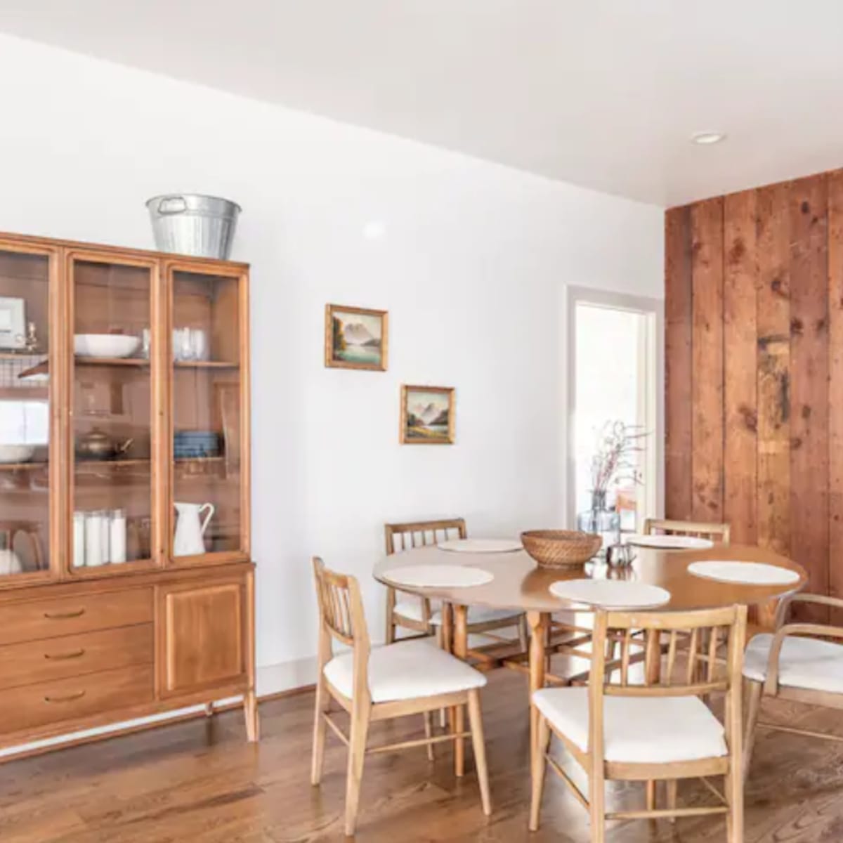 A dining area featuring a wooden table surrounded by chairs, a glass-front cabinet, and a wall made of wooden panels.