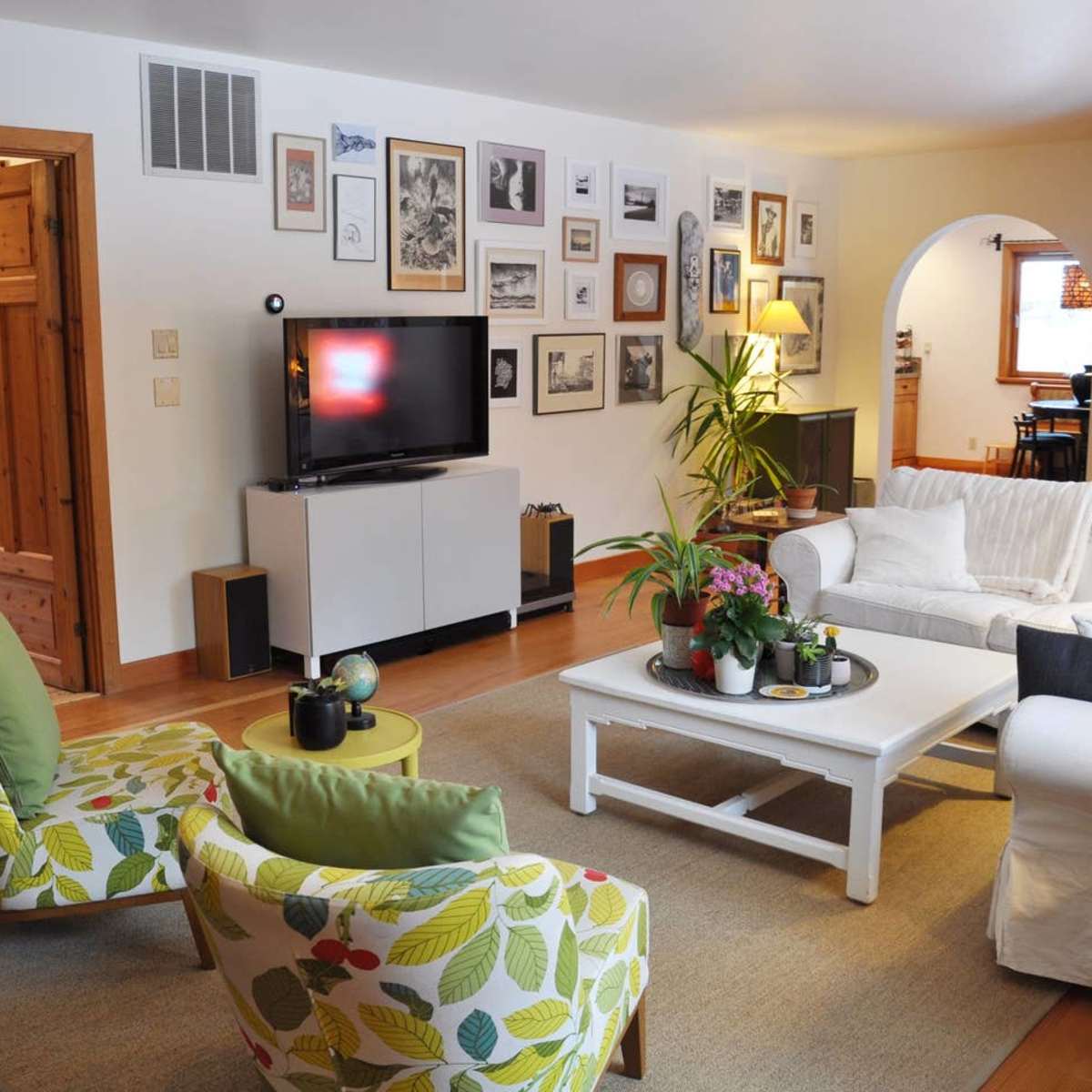 The living room features a white couch, two patterned armchairs, a coffee table with plants, and a gallery wall of framed art surrounding a television.