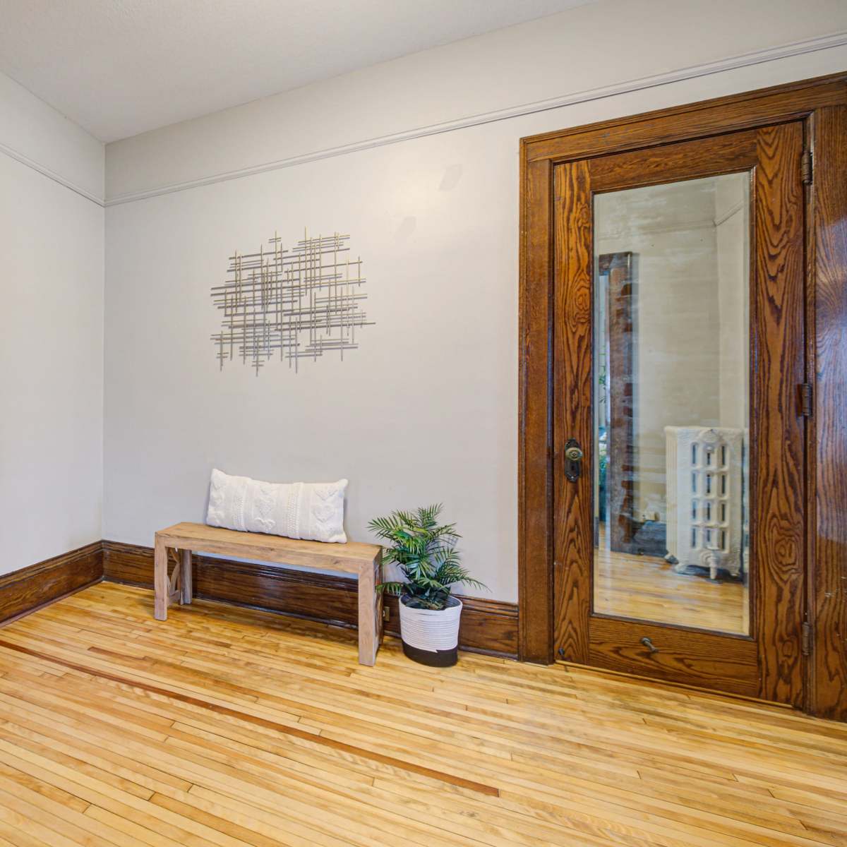 A hallway with a white door, a wooden bench, a small plant, and a mirror against light-colored walls and hardwood flooring.