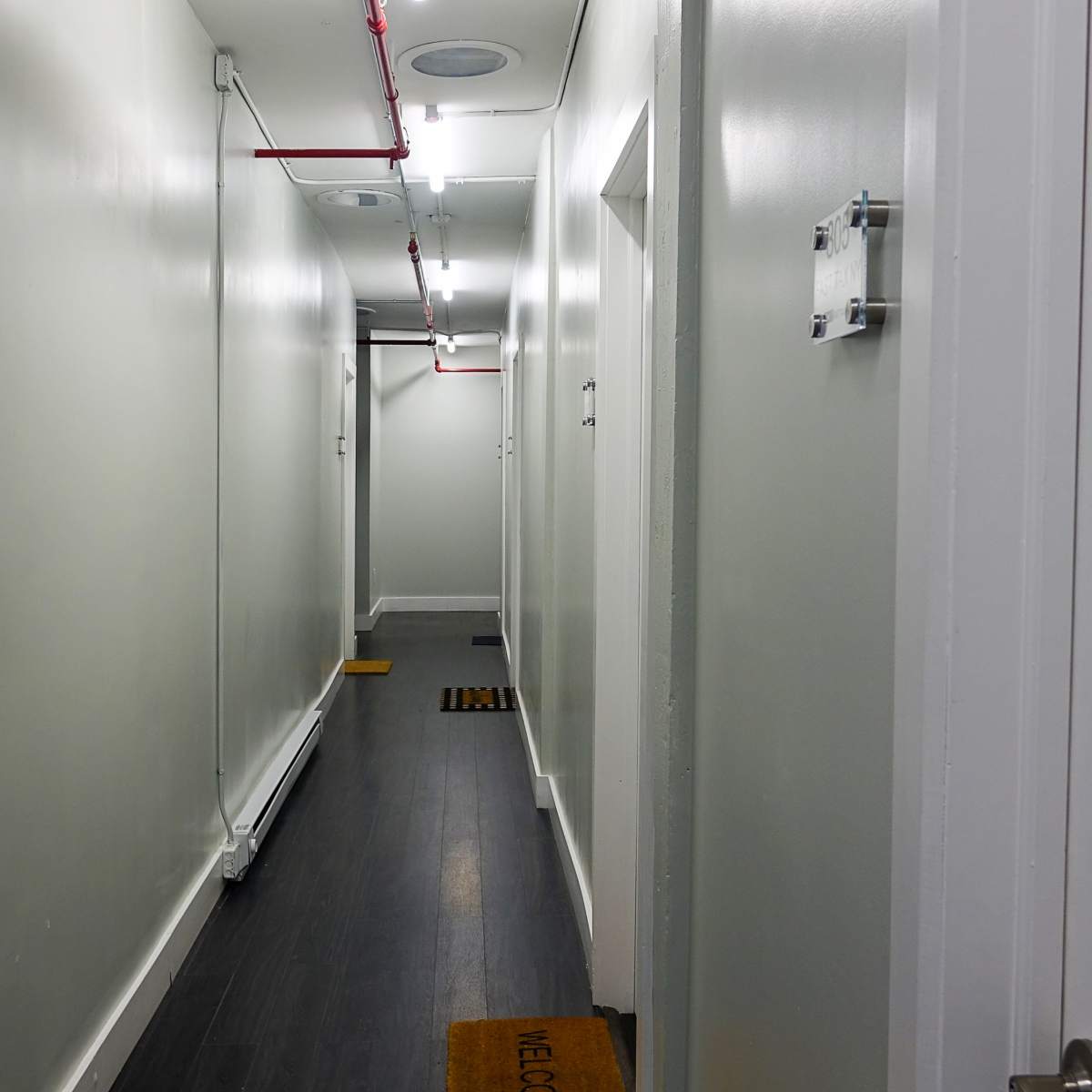 A narrow hallway with light gray walls, dark wood flooring, and several doors along each side, leading to a door at the end.