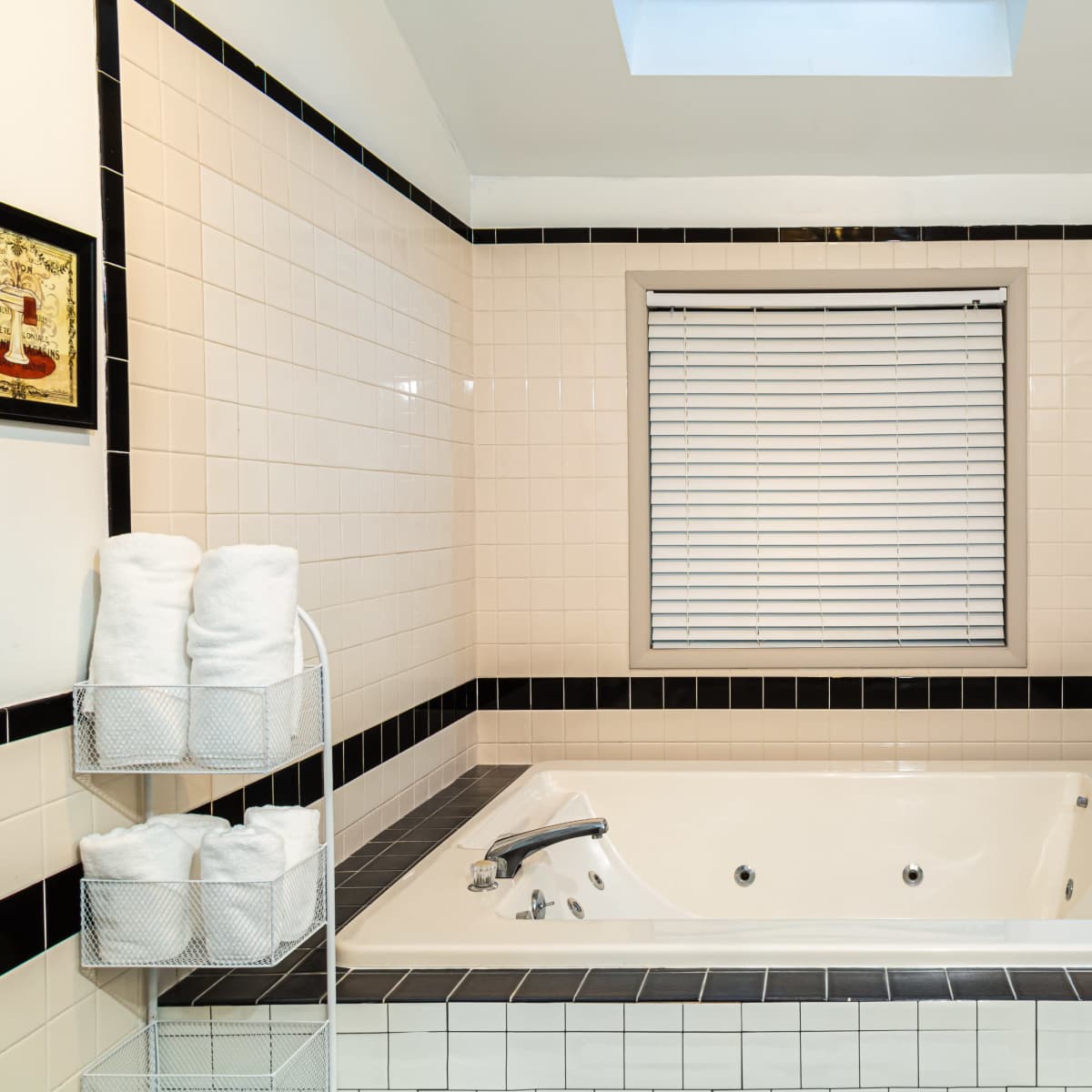 A bathroom featuring a rectangular jacuzzi tub surrounded by tiled walls, a skylight overhead, and neatly stacked white towels in a metal rack.