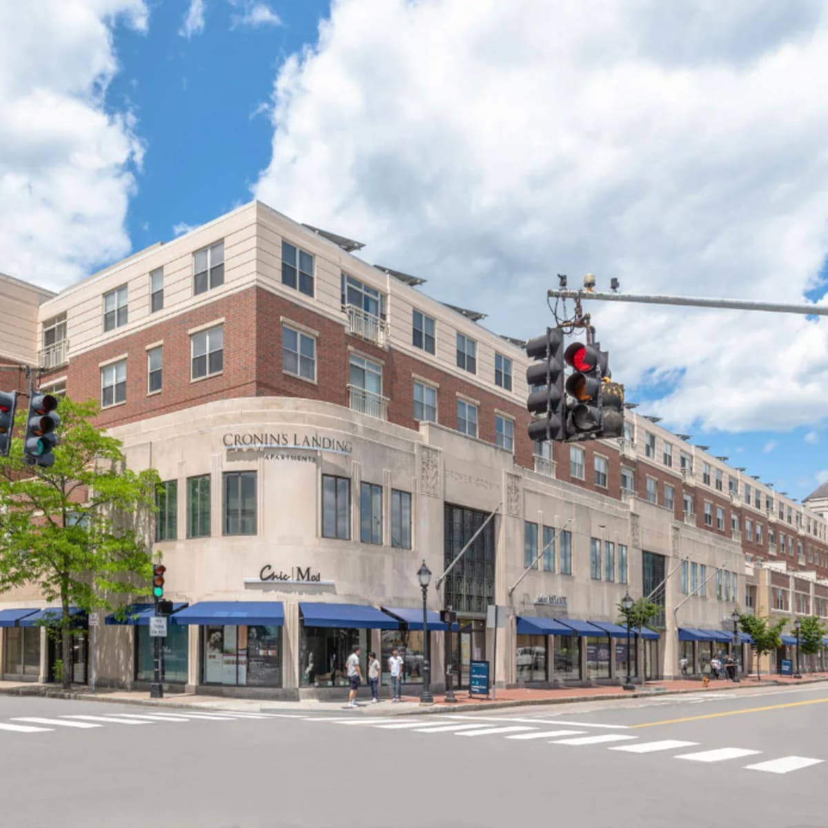 A multi-story building with retail storefronts and a traffic signal at a street intersection under a partly cloudy sky.