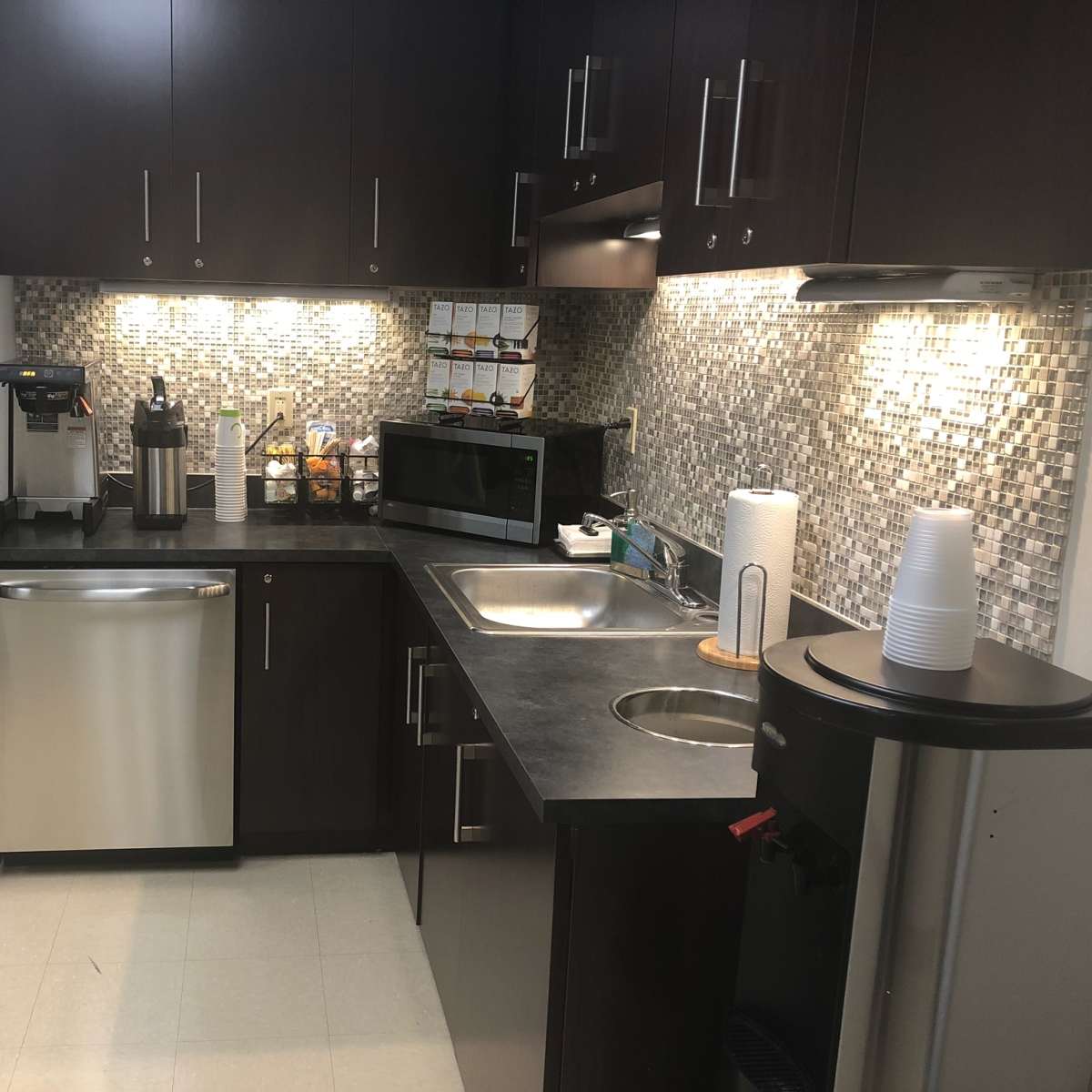A modern kitchen with stainless steel appliances, including a refrigerator and dishwasher, a microwave, and a coffee machine, set against a tiled backsplash.