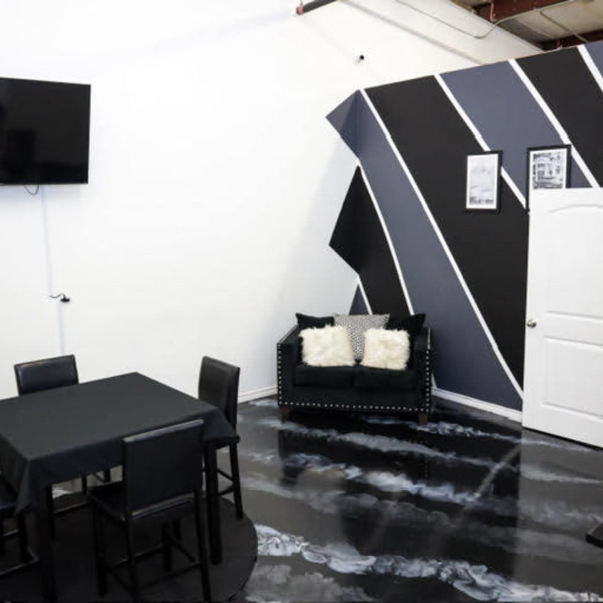 A modern room with a square black table and four chairs, a couch with cushions, a mounted television, and a painted accent wall featuring diagonal black and gray stripes.