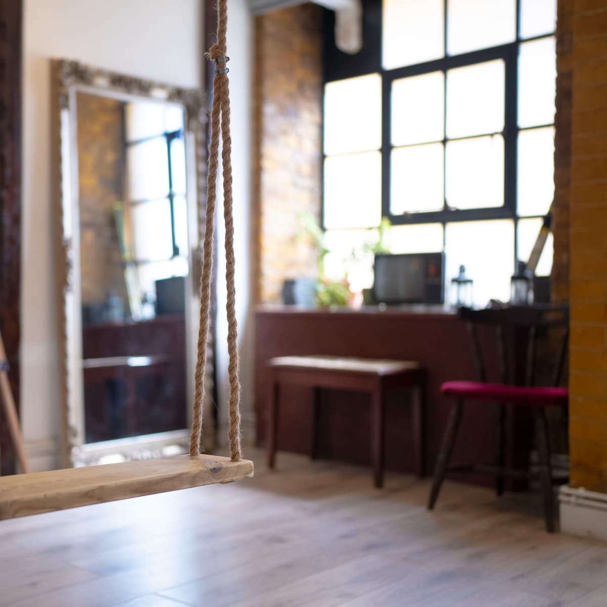 A wooden swing hanging from the ceiling in a room with large windows, a mirror, and various furniture pieces.