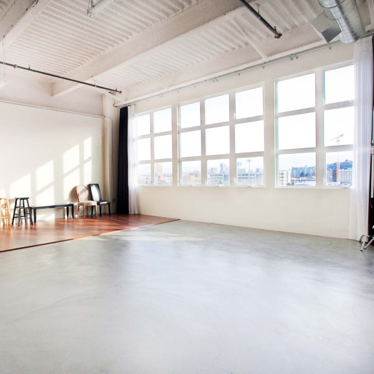 A spacious studio with large windows, wooden flooring in one section, and a few chairs positioned near the wall.
