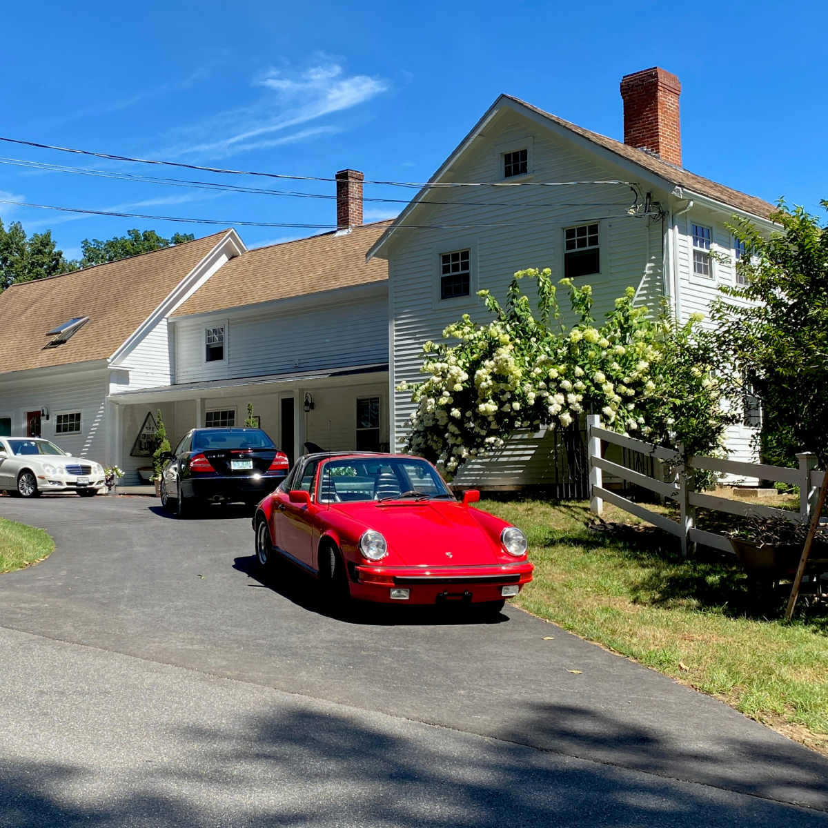 A red sports car and two other vehicles are parked in front of a large white house surrounded by greenery.