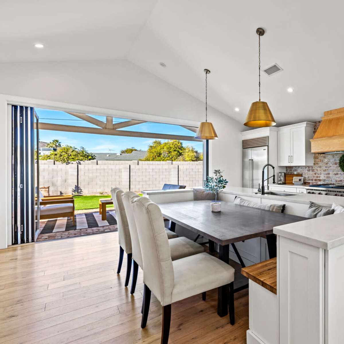 A modern kitchen features a large dining table, white cabinetry, wooden accents, and pendant lights, with a view of a backyard through sliding glass doors.