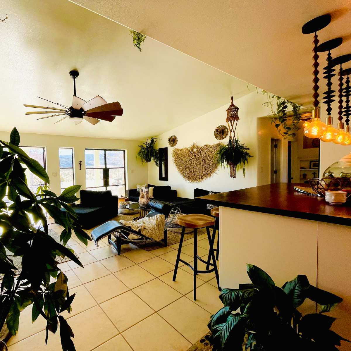 A bright living space with a ceiling fan, large windows, and a kitchen counter, surrounded by plants and brown leather furniture.