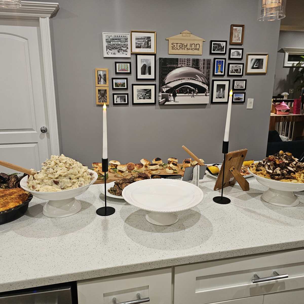 A buffet spread on a white countertop featuring various dishes, including meats, mashed potatoes, appetizers, and candle holders, with a wall of framed photos in the background.