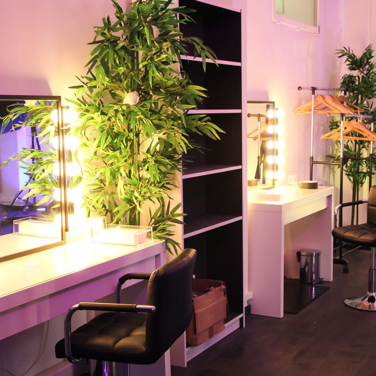 A brightly lit dressing room featuring two vanity mirrors with lights, a shelf with plants, and a black chair beside a clothing rack.