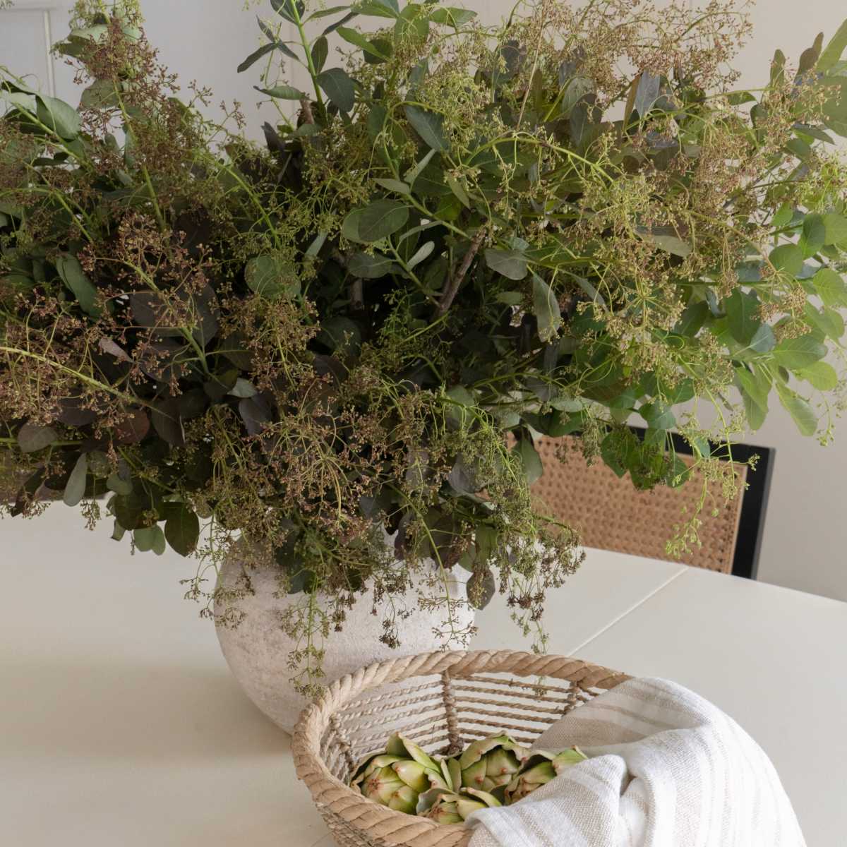 A large vase filled with greenery sits on a white table next to a woven basket containing slices of cucumber and a striped cloth.