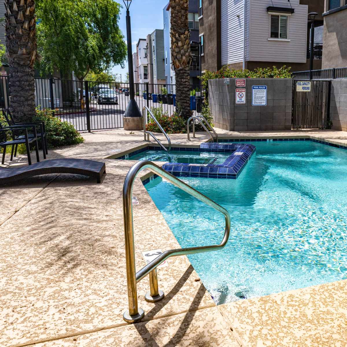 A rectangular swimming pool with a hot tub is surrounded by a concrete deck and fenced by a black gate, with lounge chairs nearby.