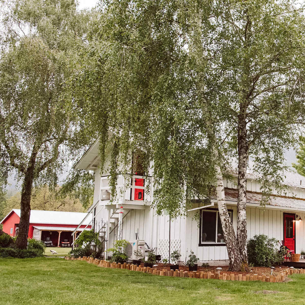 A white two-story house with red accents is surrounded by greenery and a well-maintained lawn.