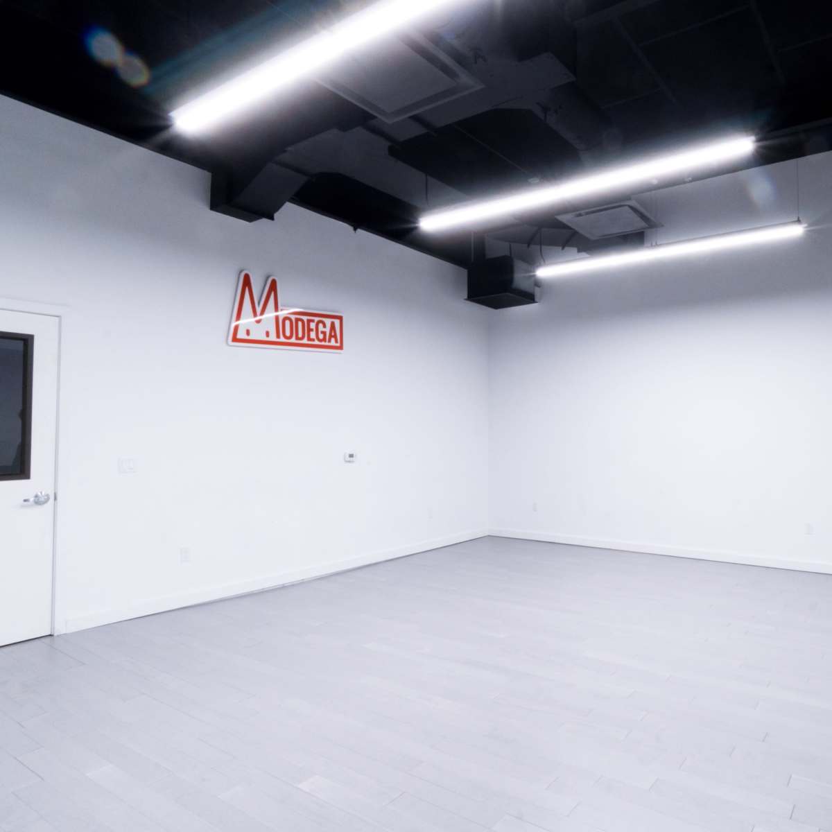 A nearly empty room with white walls, gray flooring, a door on the left, and a large mirror along the right side, featuring a neon sign that reads "MODEGA."