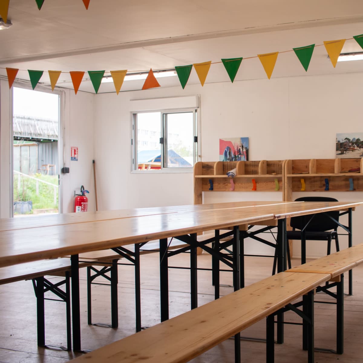 A brightly lit room features long wooden tables and benches, with colorful triangular bunting strung across the ceiling and windows.