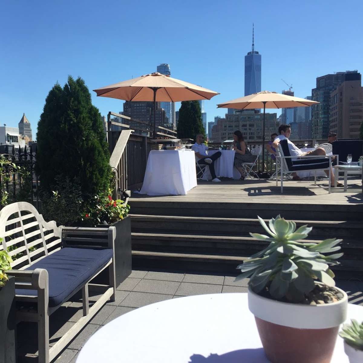 A rooftop terrace with potted plants and umbrellas, overlooking city buildings, including a tall skyscraper in the background.